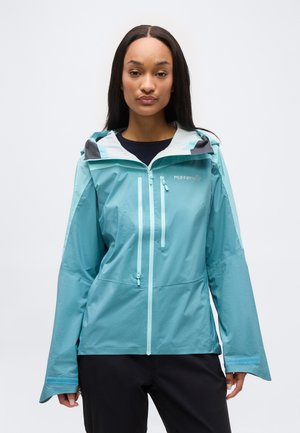 Γυναίκα με πετρόλ αδιάβροχο μπουφάν με κουκούλα, φερμουάρ στο μπροστινό μέρος και στο στήθος, στέκεται μπροστά σε ένα απλό ανοιχτόφθορο φόντο.