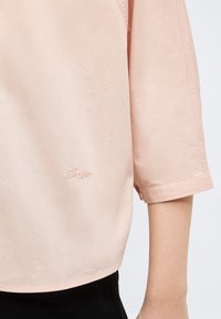 Ροζ ανοιχτό ροζ κοντομάνικο πουκάμισο με διακριτική κεντημένη υπογραφή κοντά στο τελείωμα, που φοριέται στο δεξί μπράτσο και τον κορμό πάνω σε απλό φόντο.