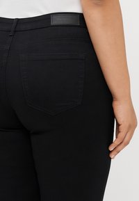 Egy személy alsó hátának és kezének közeli képén fekete, magas derekú farmernadrág látható, egyetlen hátsó zsebbel és látható márkajelzéssel.