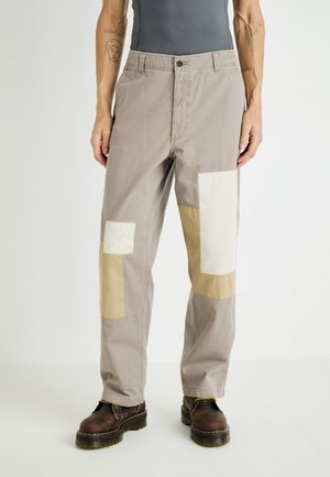 Light gray trousers with mixed fabric patches in beige and off-white. Features a relaxed fit, zip fly, and oversized pockets.