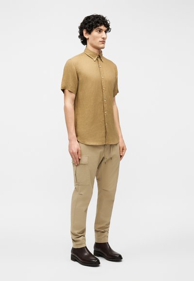 Νέος άνδρας με σγουρά μαλλιά, φοράει καφέ κοντομάνικο πουκάμισο με κουμπιά, μπεζ παντελόνι cargo και σκούρα καφέ μπότες, στέκεται πλάγια.