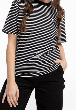 Person med flettet hår iført en sort- og hvidstribet T-shirt og sorte bukser, den ene hånd i lommen og den anden hvilende ved siden.