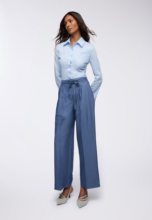Camicia azzurro chiaro abbinata a pantaloni blue in stile wide-leg con vita alta. Presenta un cinturino in vita e scarpe con un leggero motivo.