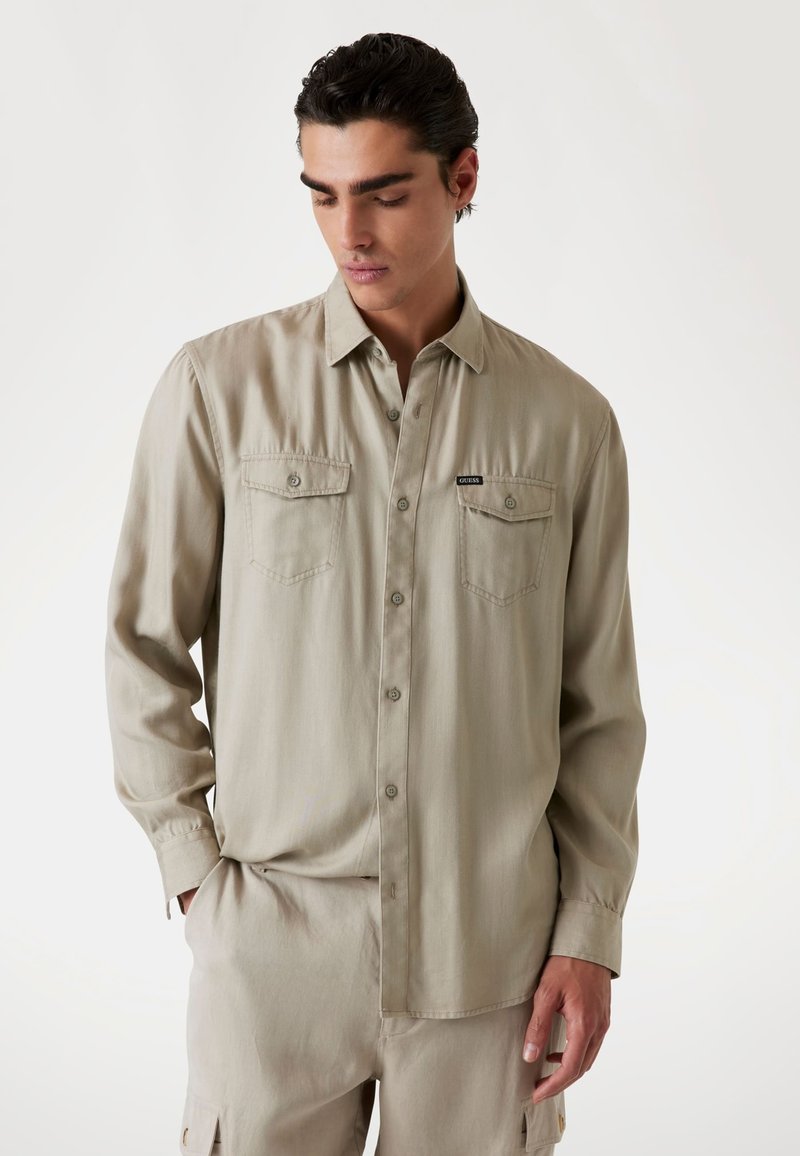 Camisa de manga larga color beige claro con un cierre de botones en el frente, dos bolsillos en el pecho y una textura sutil. Presenta un pequeño logo negro en el bolsillo izquierdo.