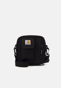 Borsa a tracolla Carhartt nera con tasca frontale, tasca con zip, tracolla regolabile e patch del logo su sfondo bianco semplice.