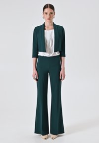 Completo pantaloni verde su misura con giacca corta, camicetta bianca sotto, tessuto texturizzato, pantaloni svasati e scarpe beige a punta.
