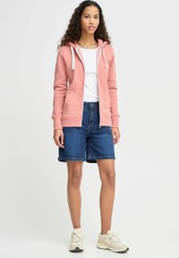 Ružová mikina na zips s bielymi šnúrkami, v kombinácii s modrými džínsovými šortkami. Model má na sebe béžové tenisky a stojí na neutrálnom pozadí.