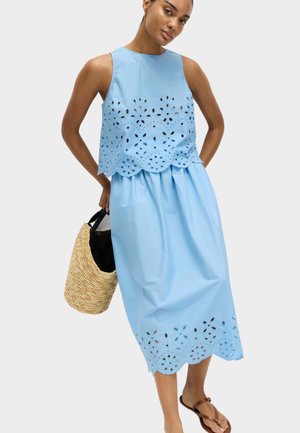 Kvinde iført en ærmeløs lyseblå kjole med blomsterudskæringer, der holder en stråtaske og bærer brune sandaler.
