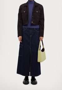 Zwarte cropped jacket met gouden knopen, paarse geribbelde coltrui, lange donkere denim rok met een split aan de voorkant, zwarte schoenen en een gele handtas.
