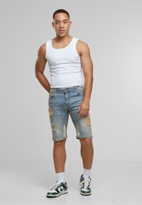 Traperice kratke hlače svjetloplave boje s istrošenim detaljima i izblijedjelim efektom. Uparene s bijelom majicom bez rukava i zelenim Nike tenisicama, naslonjene na sivu pozadinu.