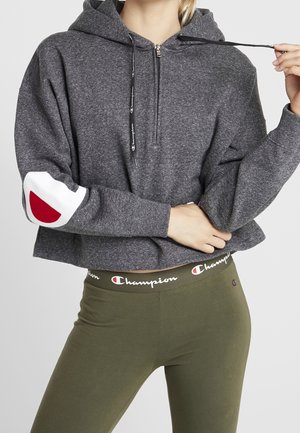 Personne portant un sweat à capuche court gris avec une fermeture éclair frontale et un logo sur la manche, associé à un legging vert olive avec une ceinture à logo.