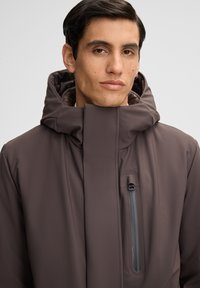 Junger Mann mit kurzen dunklen Haaren, der eine dunkelbraune Jacke mit hohem Kragen und einer vertikalen Reißverschlusstasche auf der Brust trägt, vor einem neutralen Hintergrund.