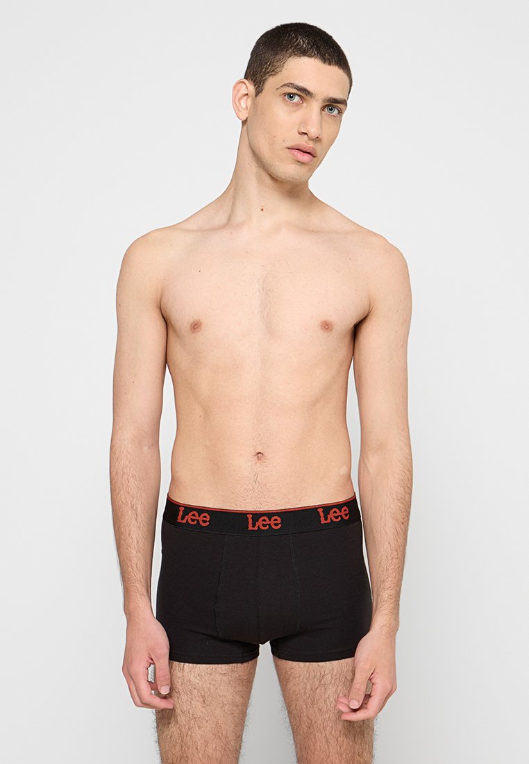 Un modèle masculin se tenant contre un fond uni, portant un boxer noir de marque Lee avec une ceinture logo rouge, regardant légèrement sur le côté.