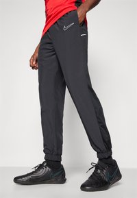 Schwarze Nike-Hosen mit elastischen Bündchen, Seitentaschen und einem weißen Logo auf dem Oberschenkel. Getragen mit schwarzen Sportschuhen mit türkisfarbenen Akzenten.