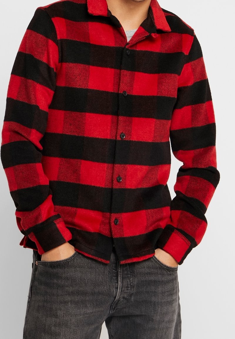 Röd och svart rutigt flanellskjorta med knapplist, långa ärmar, en krage och en mjuk textur, bärs över grå jeans.