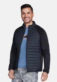 Marineblauwe jas met een gequiltte voorkant, zwarte mouwen en een hoge kraag. Voorzien van een ritssluiting en een gestructureerde stof. Draagt over een blauwe shirt.