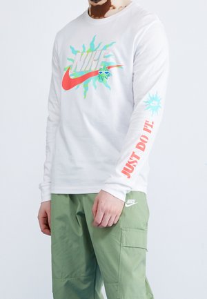 Biała koszula z długim rękawem z kolorowym logo Nike i grafiką; na rękawie czerwony napis "JUST DO IT!"; w połączeniu z zielonymi spodniami typu cargo.