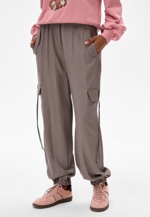 Osoba nosząca szare joggery cargo ze ściągaczami, z rękami w kieszeniach, w połączeniu z różowymi trampkami i różową bluzą w kwiaty.