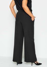 Pantaloni neri a gamba larga con vita elasticizzata, tasche laterali e una texture liscia, abbinati a tacchi neri. Design semplice e funzionale.