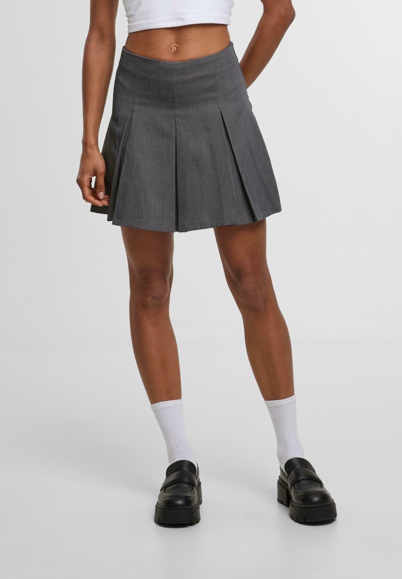 Harmaa pleatattu hame, jossa on istuva vyötärö, sileä rakenne ja polvipituinen muotoilu, yhdistettynä mustiin platform-korkoihin ja valkoisiin sukkiin.