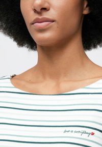 Κοντινό πλάνο ενός ατόμου που φοράει ένα άσπρο και τιρκουάζ ριγέ πουκάμισο με την ένδειξη "η αγάπη είναι τα πάντα" και μια μικρή κόκκινη καρδιά κεντημένη κοντά στη κλείδα.