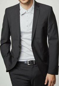 Veste de costume noire ajustée sur un polo gris clair à motif texturé, associée à un pantalon noir et une boucle de ceinture argentée.