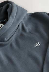 Graues, weiches Langarmoberteil mit hohem Kragen; enthält ein weißes, gesticktes Logo mit der Aufschrift "self." nahe dem Saum. Sanfte Textur, lässiges Design.
