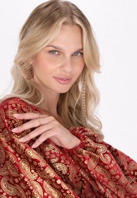 Roter Stoff mit goldfarbenem Paisleymuster; das Modell trägt goldene Ohrringe und einen zarten Ring und präsentiert dabei strukturierte, fließende Ärmel.