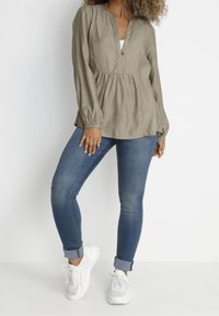Kaki langærmet bluse med V-udskæring og knapdetaljer, parret med tætsiddende blå jeans og hvide sneakers. Blødt, struktureret stof.