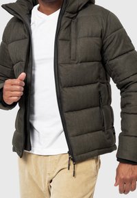 Nafukaná tmavě zelená bunda s kapucí, prošívaný design, zapínání na zip a boční kapsy. Nositá přes bílé tričko a světlé kalhoty.