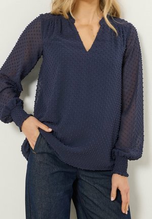Kvinde iført en mørkeblå tekstureret bluse med lange, gennemsigtige ærmer og smockede manchetter, kombineret med mørkeblå jeans, hånd i lommen.