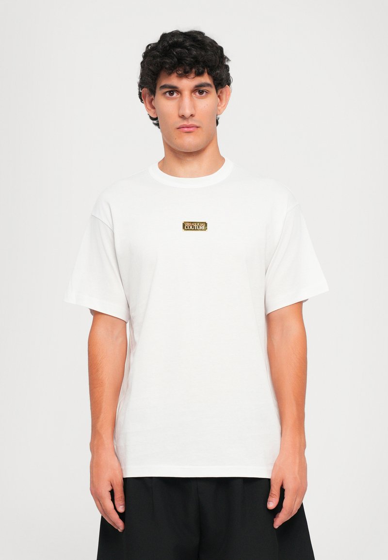 Λευκό βαμβακερό μπλουζάκι με κοντά μανίκια, που διαθέτει ένα ορθογώνιο χρυσό λογότυπο στο στήθος. Απλός σχεδιασμός με λαιμόκοψη.