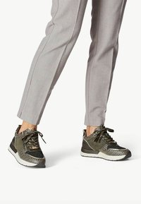 Sneakers atletici verdi con accenti texturizzati, lati a fantasia e chiusura con lacci, abbinati a pantaloni grigi su misura.