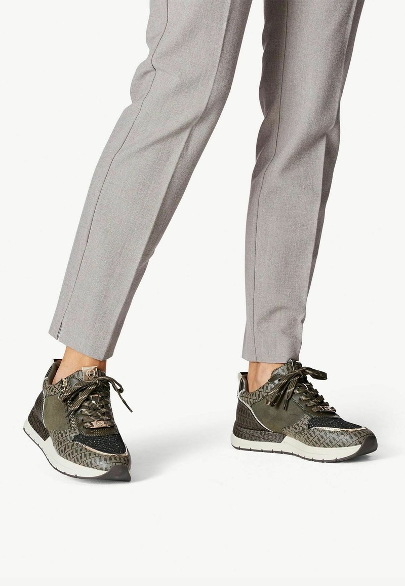 Sneakers atletici verdi con accenti texturizzati, lati a fantasia e chiusura con lacci, abbinati a pantaloni grigi su misura.