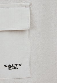 Bežna tkanina s teksturiranim mrežastim vzorcem, ki ima zgornji žep s poklopcem in črn logotip z napisom "SALTY D.G." v spodnjem kotu.