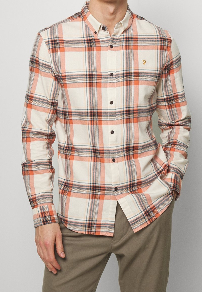 Homme portant une chemise à carreaux boutonnée à manches longues orange, blanche et grise, associée à un pantalon beige, main dans la poche.