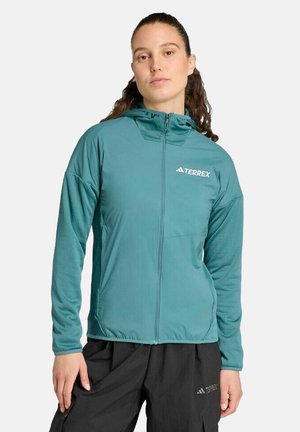 Femme portant une veste zippée Terrex turquoise avec capuche et un pantalon Terrex noir, debout devant un fond clair uni.