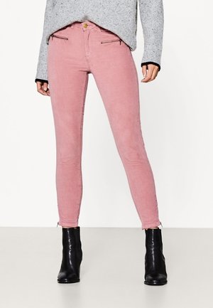 Różowe spodnie ze sztruksu o dopasowanym kroju, z kieszeniami na suwak z przodu, noszone z szarym swetrem i czarnymi botkami na tle jednolitego tła.