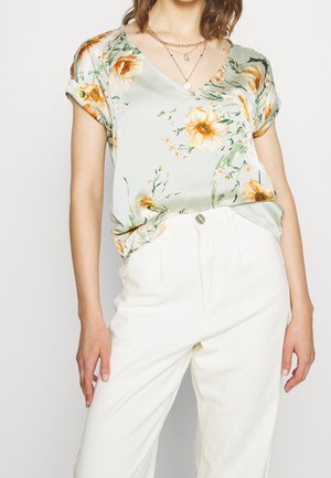 Kobieta nosząca jasną zieloną bluzkę w kwiaty z krótkim rękawem włożoną do białych spodni z wysokim stanem z warstwowymi złotymi naszyjnikami.