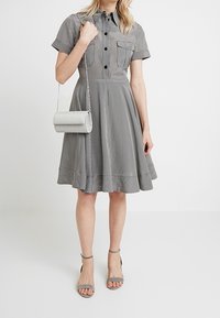 Femme portant une robe grise à manches courtes à carreaux avec boutons sur le devant et poche poitrine, tenant un sac à chaîne argentée, portant des talons gris à brides à la cheville.