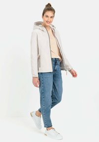Lette, beige jakke med hætte, lynlås lukning og sidelommer. Båret over en stribet skjorte og parret med blå jeans og hvide sneakers.