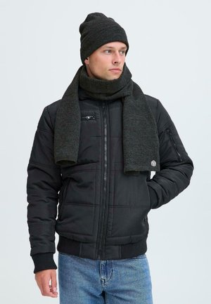 Uomo che indossa un berretto nero, una sciarpa grigio scuro, una giacca nera con zip e jeans blu, che guarda alla sua sinistra su uno sfondo chiaro.