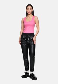 Woman wearing a sleeveless pink top, black faux leather jogger pants, white socks, and black chunky shoes standing on a plain background.