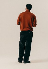 Άνδρας με κοντά σγουρά μαλλιά, φορώντας γυαλιά, καφέ πουλόβερ, μαύρο παντελόνι και μαύρα παπούτσια, στέκεται γυρισμένος προς τα πίσω μπροστά σε ένα απλό φόντο.