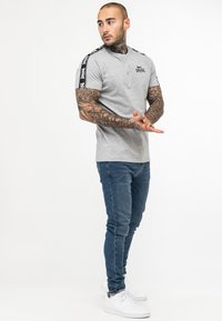 Tricou gri cu mânecă scurtă, cu dungi laterale negre, având un logo al brandului pe piept. Purtați cu blugi albaștri și adidași albi.
