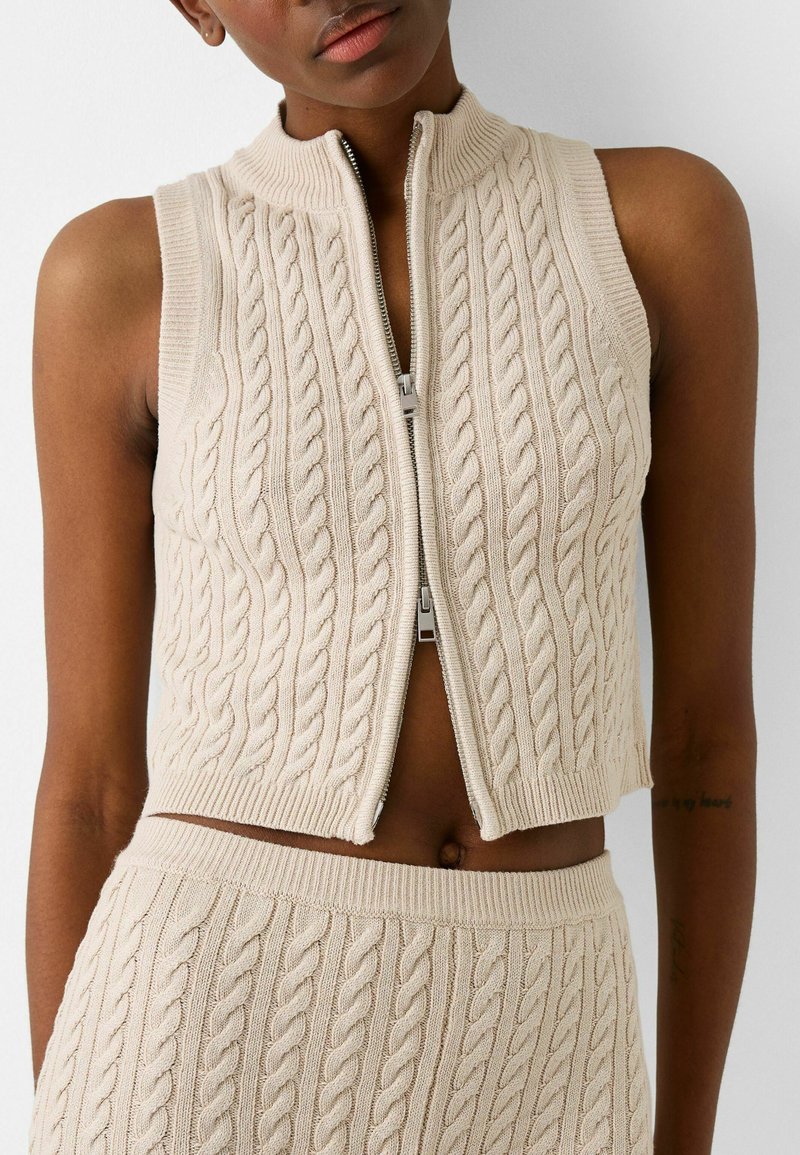 Gilet beige in maglia con zip, motivo a trecce, colletto alto e design senza maniche, abbinato a un pantalone a trecce coordinato.