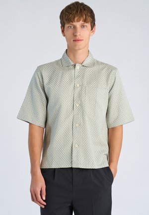 Junger Mann, der ein helles kurzärmliges Hemd mit Muster und schwarze Hose trägt, steht mit einer Hand in der Tasche vor einem schlichten Hintergrund.