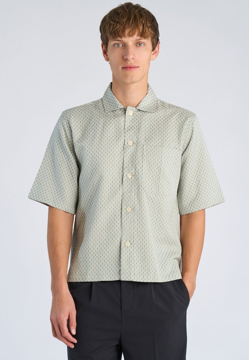 Νέος άνδρας που φοράει ένα ανοιχτόχρωμο πουκάμισο με κοντά μανίκια και σχέδιο, και μαύρο παντελόνι, στέκεται με το ένα χέρι στην τσέπη, μπροστά σε απλό φόντο.