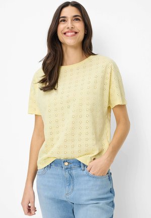 Donna sorridente che indossa una blusa gialla chiaro a maniche corte con occhielli, infilata in jeans azzurri, con una mano in tasca, su sfondo bianco.