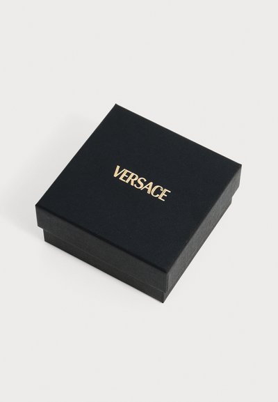 Μαύρο τετράγωνο κουτί με χρυσό λογότυπο Versace στο καπάκι, τοποθετημένο σε λευκή επιφάνεια.